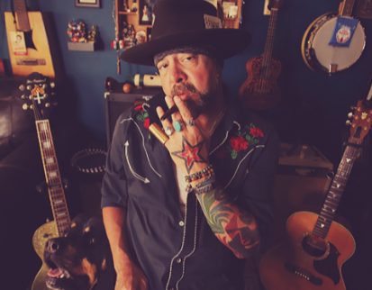 Daniel “Danny” Carbone in a black wide-brim hat and embroidered western shirt, guitars and a banjo on the blue wall behind him, his dog at his side